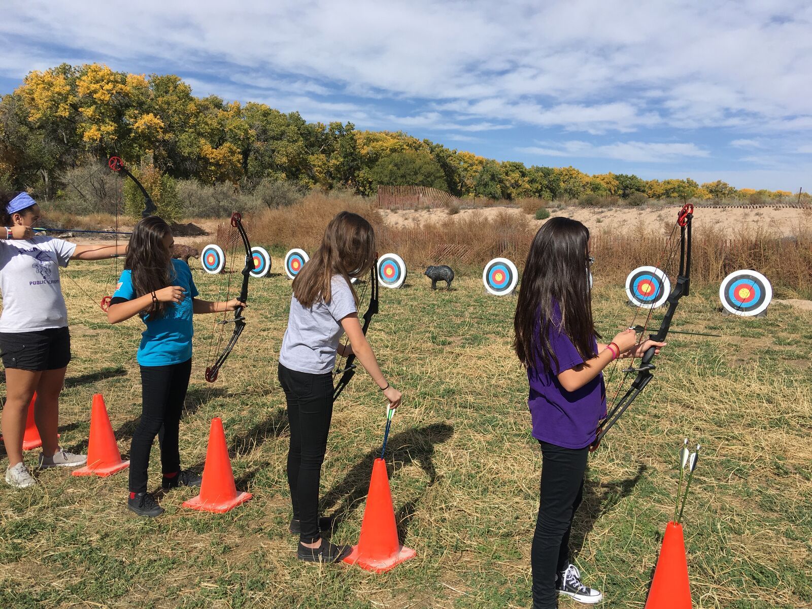 Three girls draw their bows at a line of targets, cottonwoods turning gold behind them.