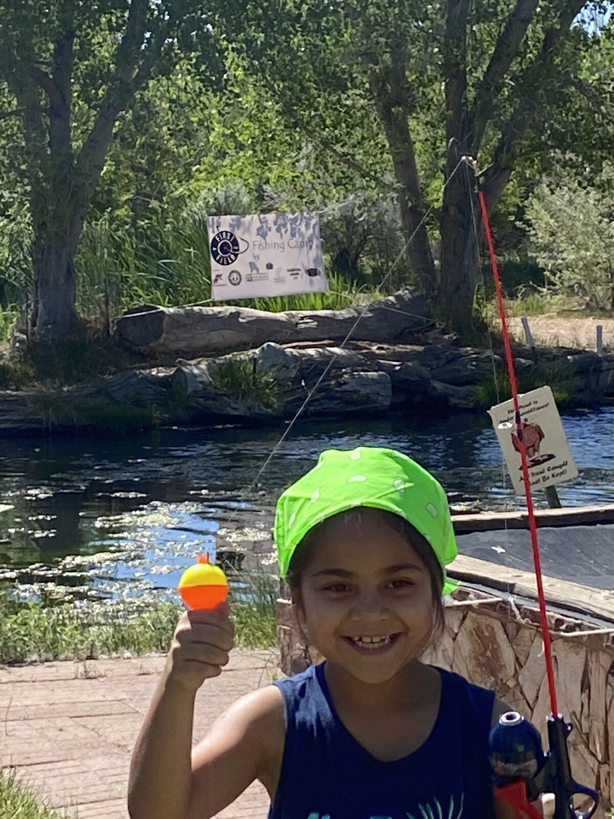 A grinning kid in a green bandana holds up an orange bobber beside the pond.