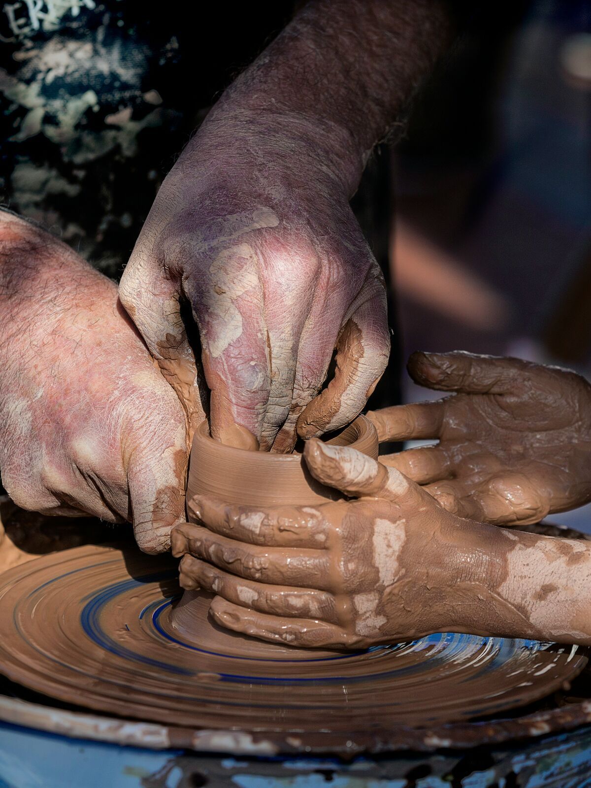 A potter's hands guide a child's as they shape a clay pot on the wheel.