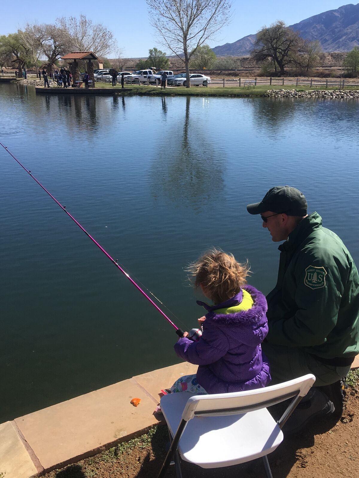 A child in a purple jacket fishes from the edge of a pond beside a ranger.