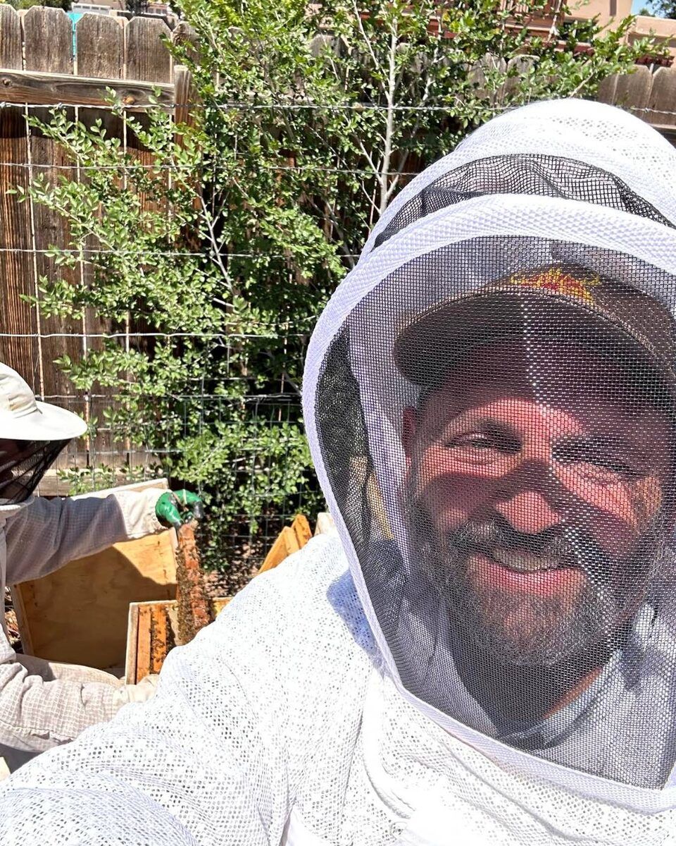 Yancey smiles through a beekeeper’s veil, a frame of bees lifted from the hive behind him.