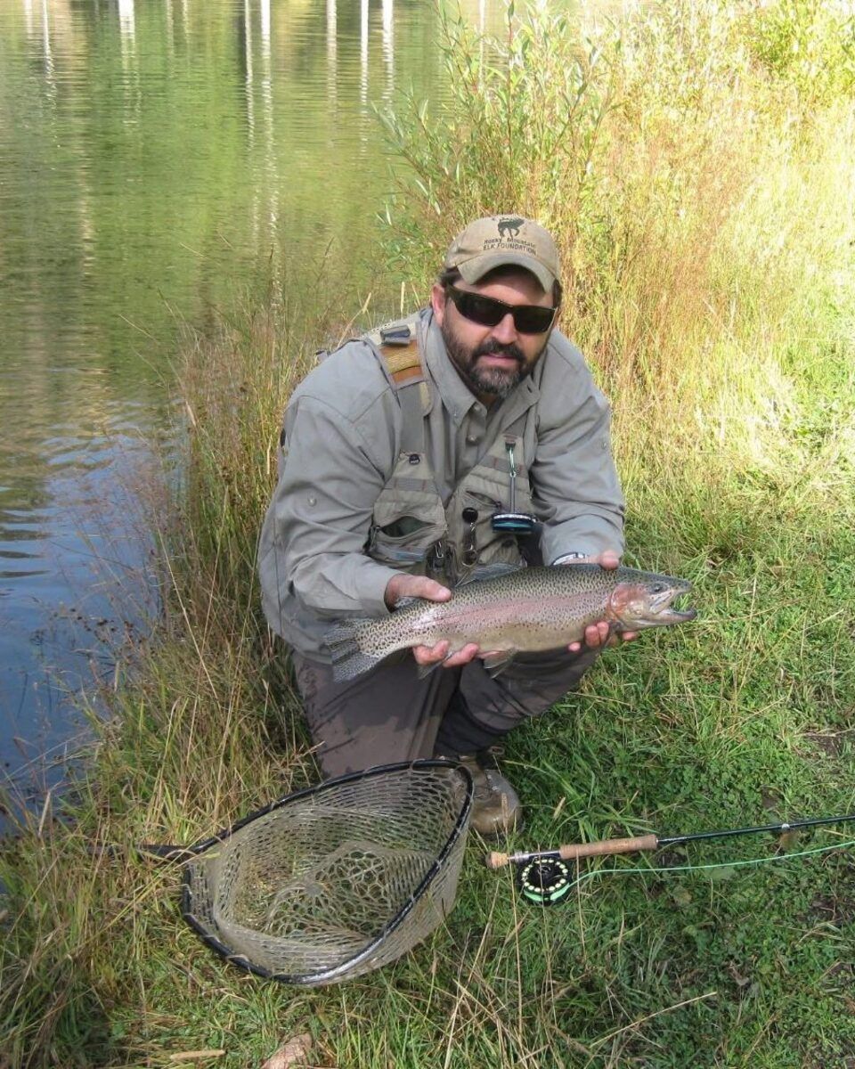 Yancey crouches at the edge of a lake with a trout, a net and a fly rod in the grass beside him.