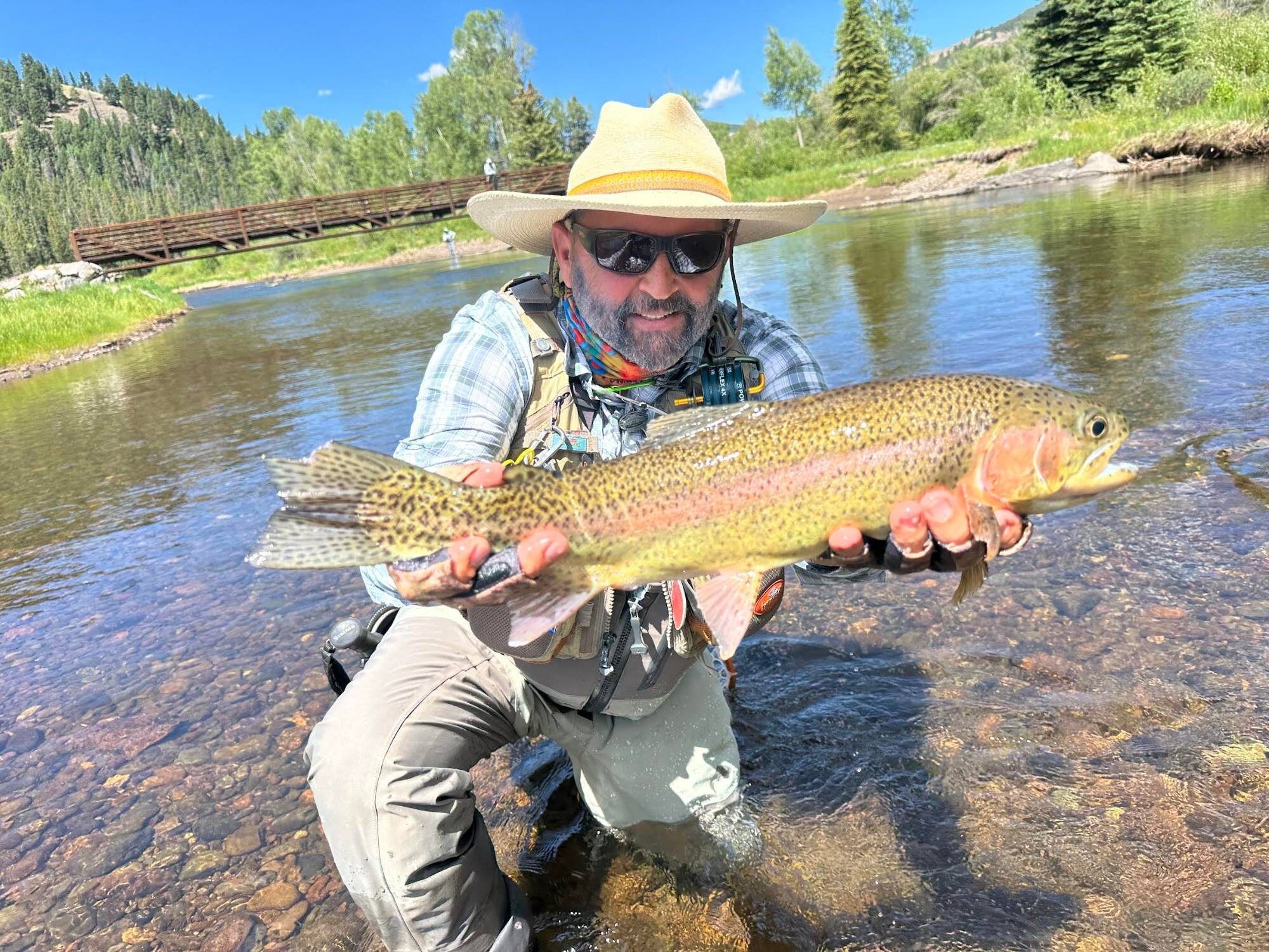 Yancey Ranspot, in a straw hat and sunglasses, kneels in a clear river and holds up a big rainbow trout.