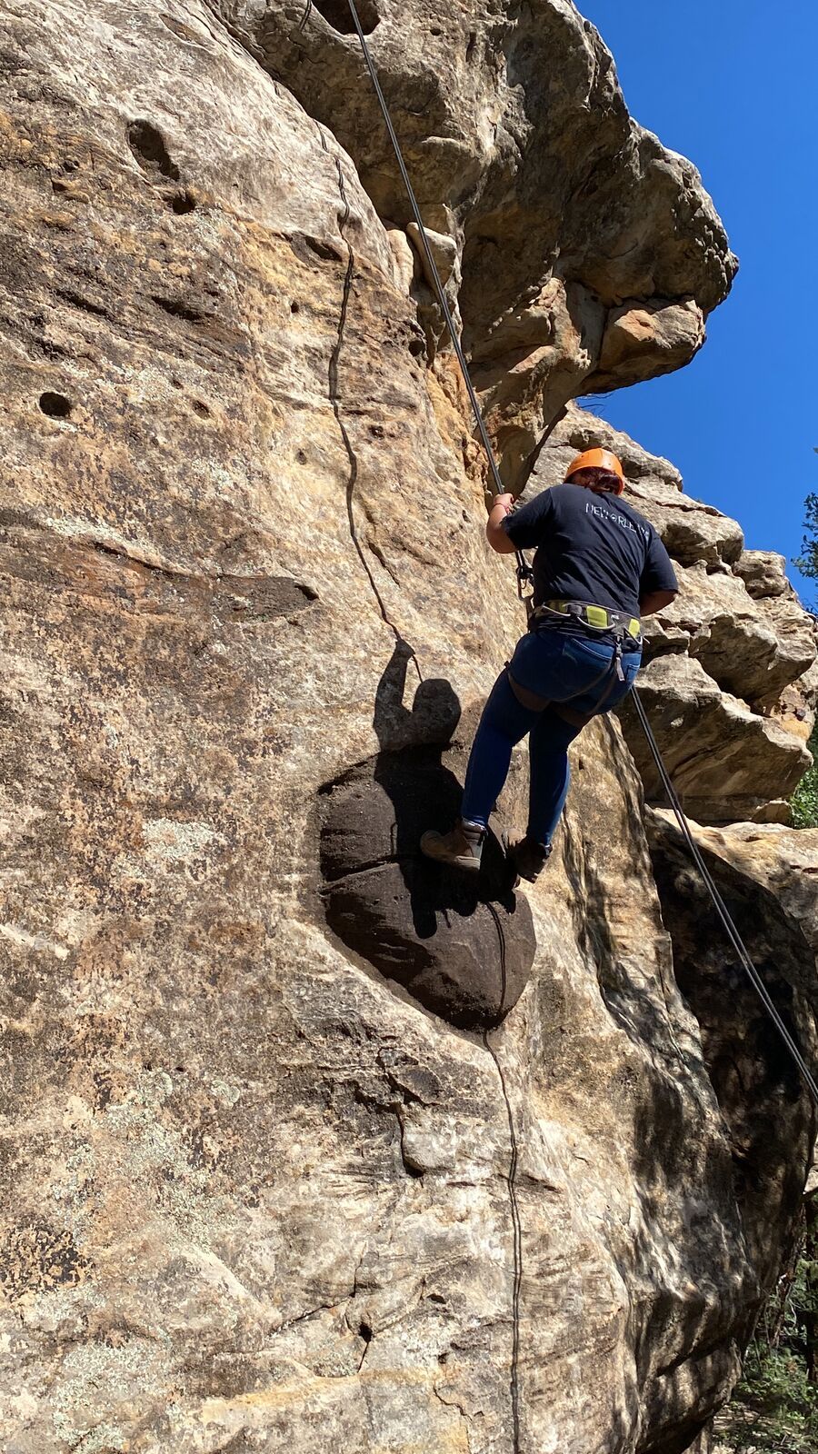 A climber in an orange helmet and harness works up a pocketed sandstone face on a rope, back to the camera, under a deep blue sky.