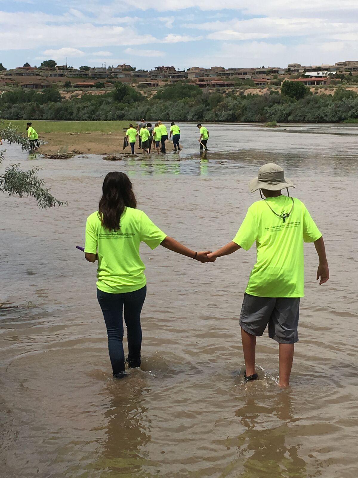 Two people in yellow shirts walk hand in hand into the river, seen from behind.