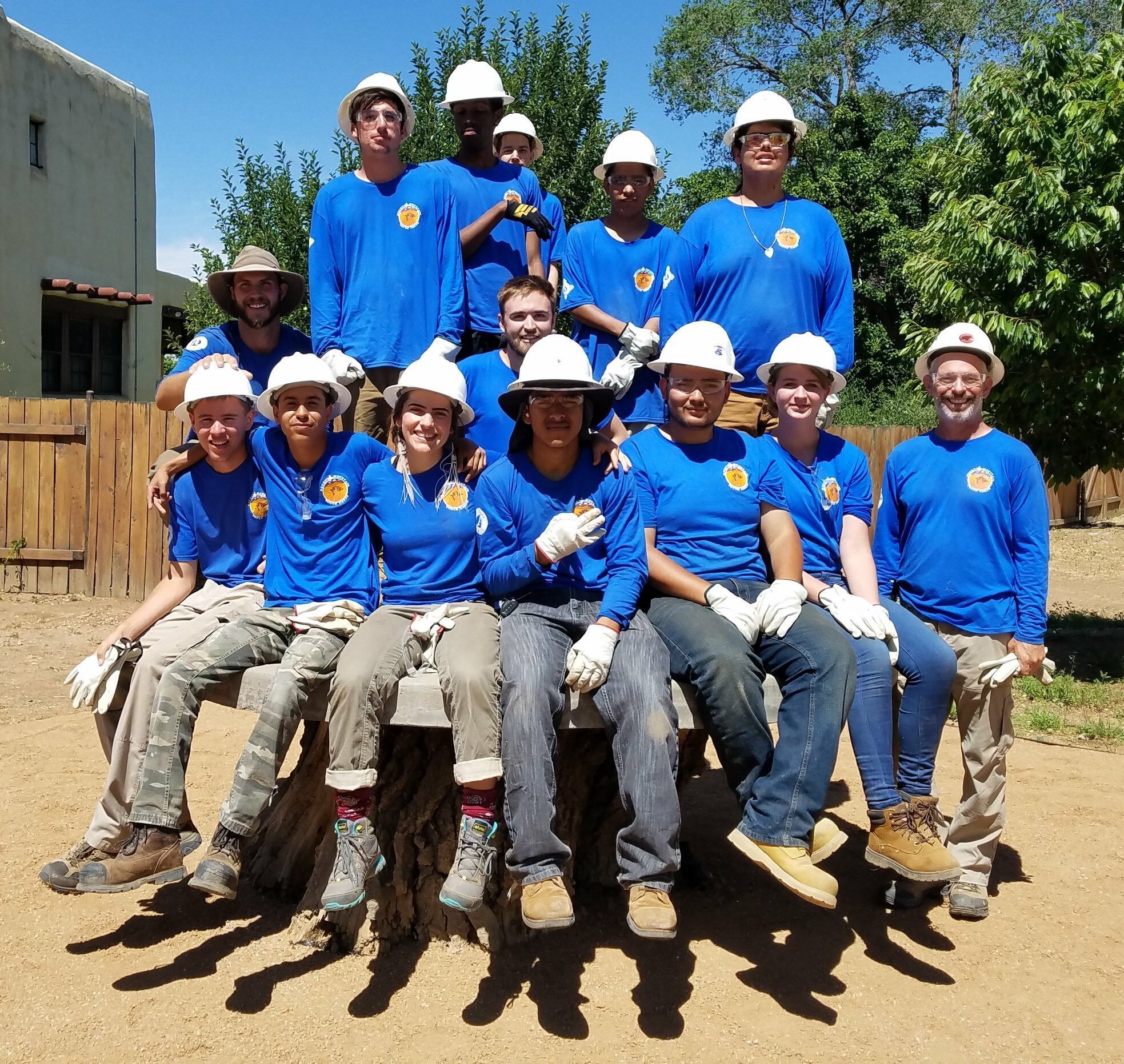 A crew in matching blue shirts and white hard hats gathers on and around a log bench, work gloves on.