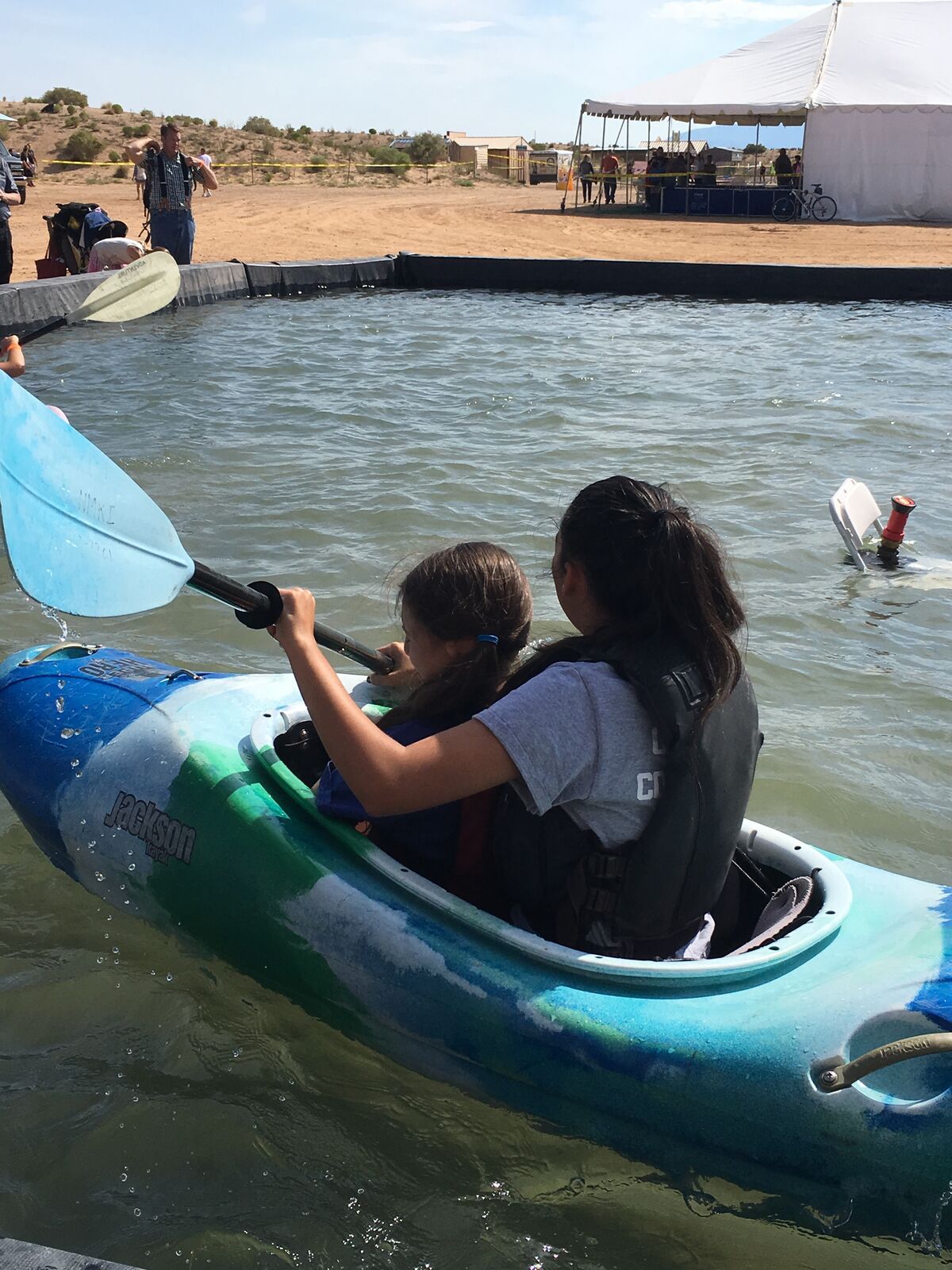 Two girls in life jackets share a blue kayak, one pulling hard on the paddle.