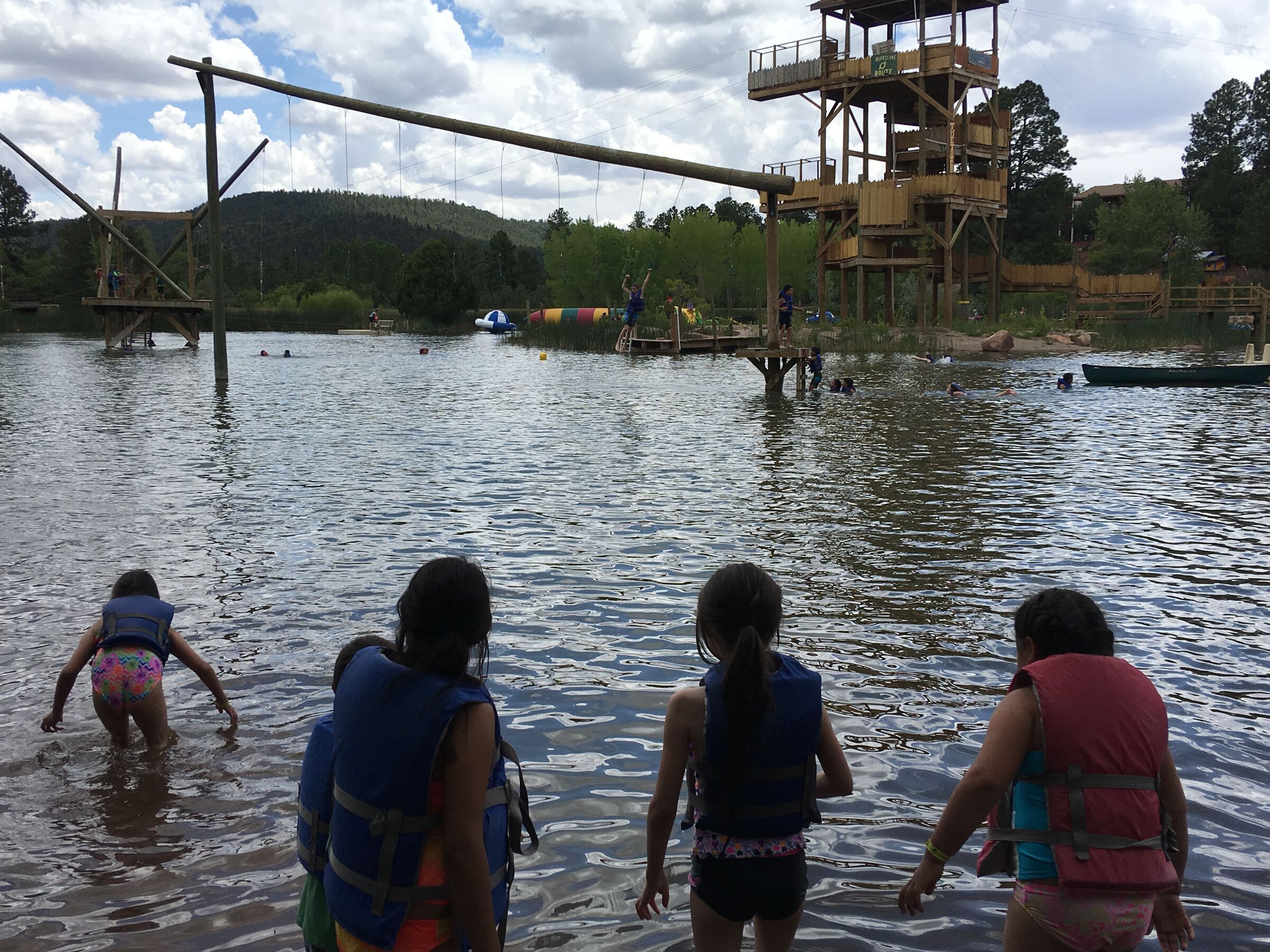 Kids in life jackets wade into the lake at Glorieta, seen from behind.