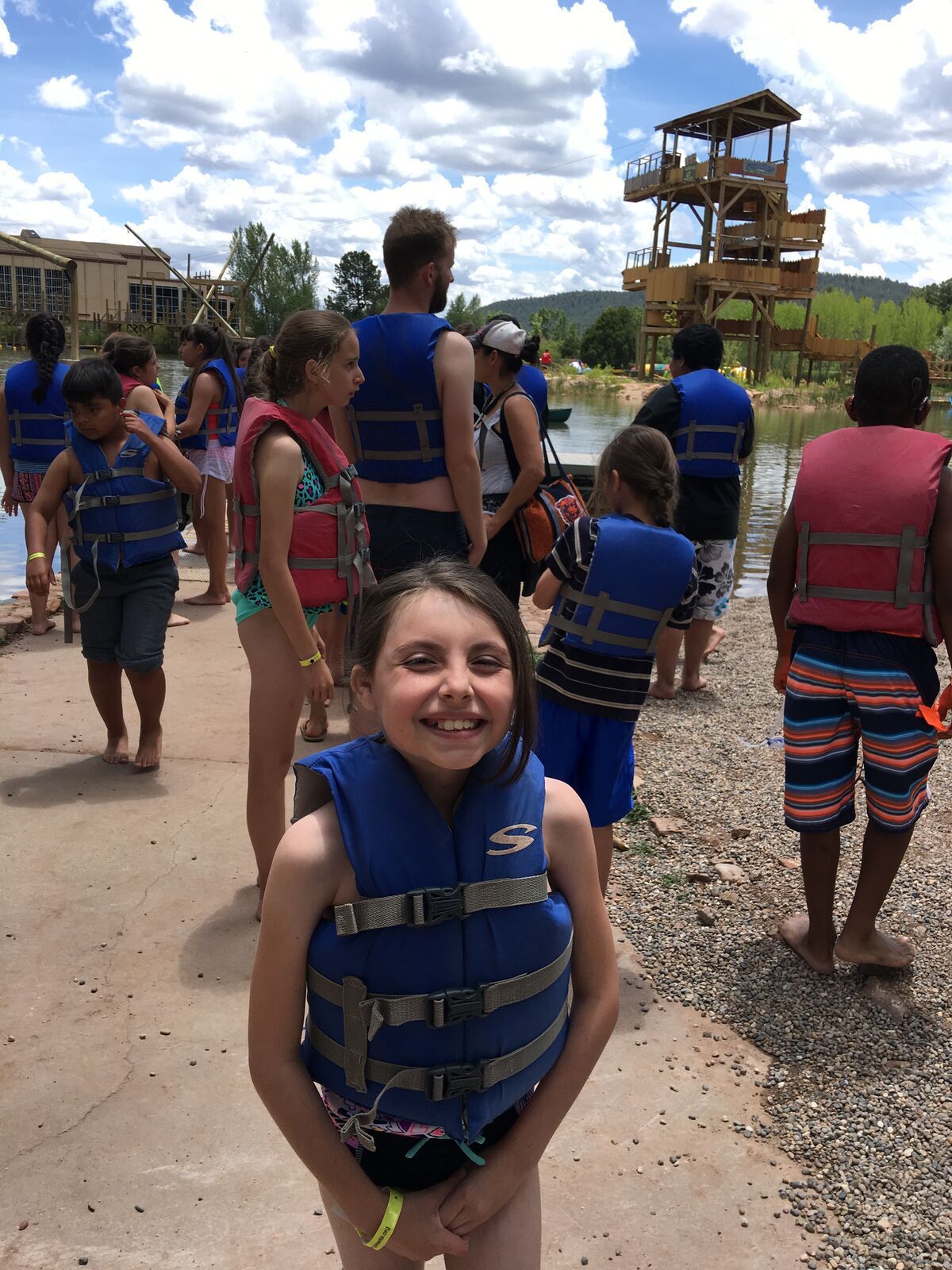 A grinning girl in a blue life jacket on the lake beach at Glorieta.