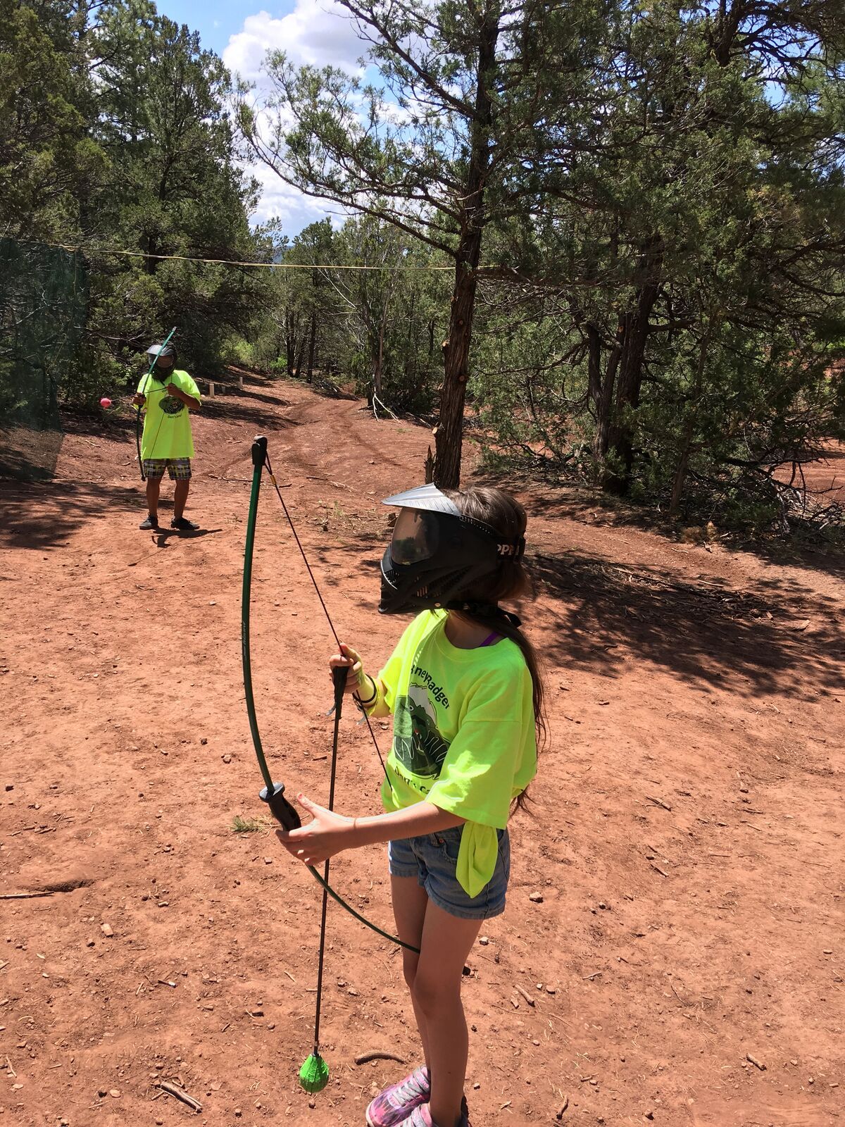 A girl in a bright green shirt holds her bow on a red-dirt range among ponderosa pines.