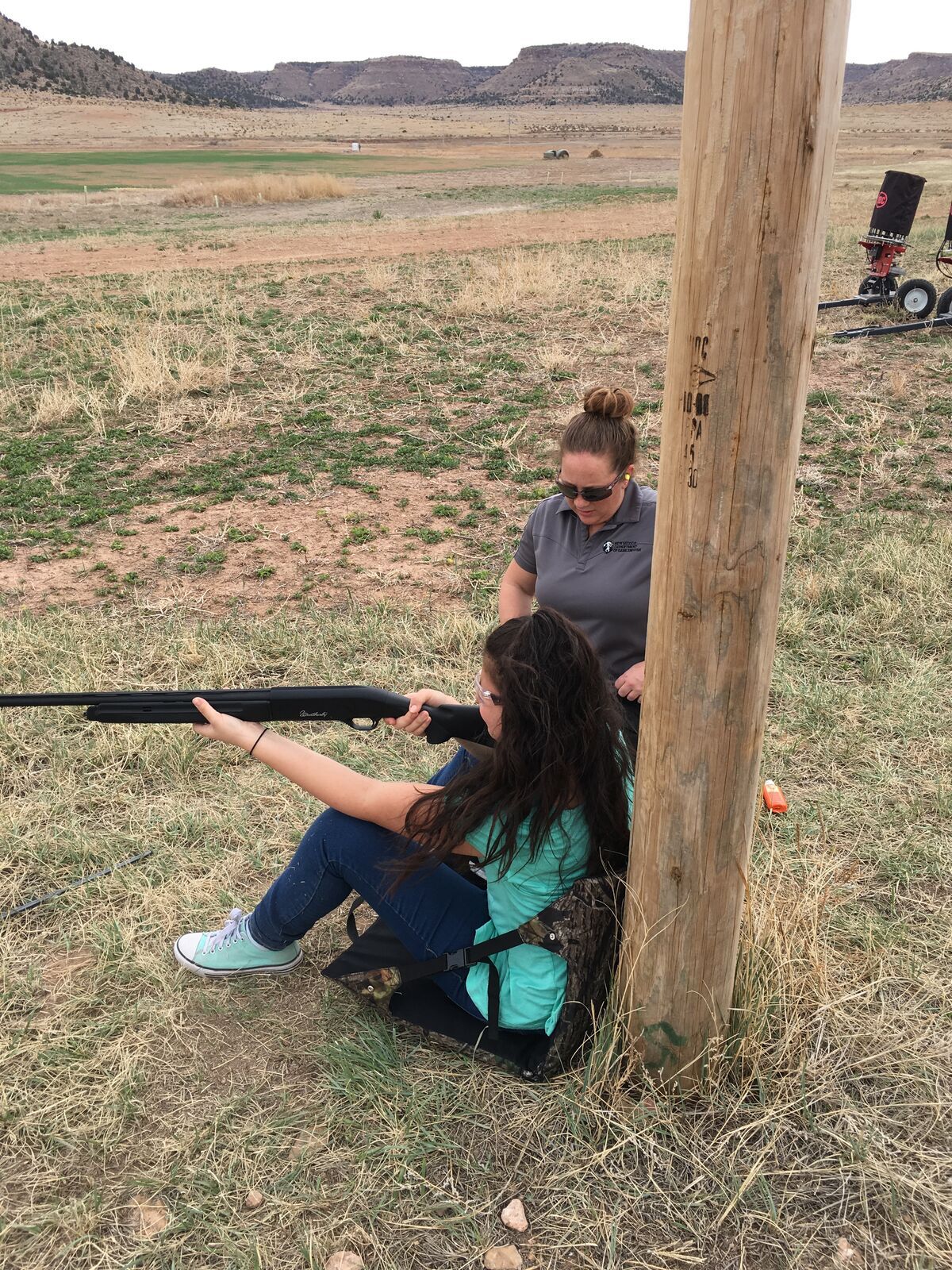 A girl sits braced against a post and aims a shotgun downrange, an instructor standing beside her.