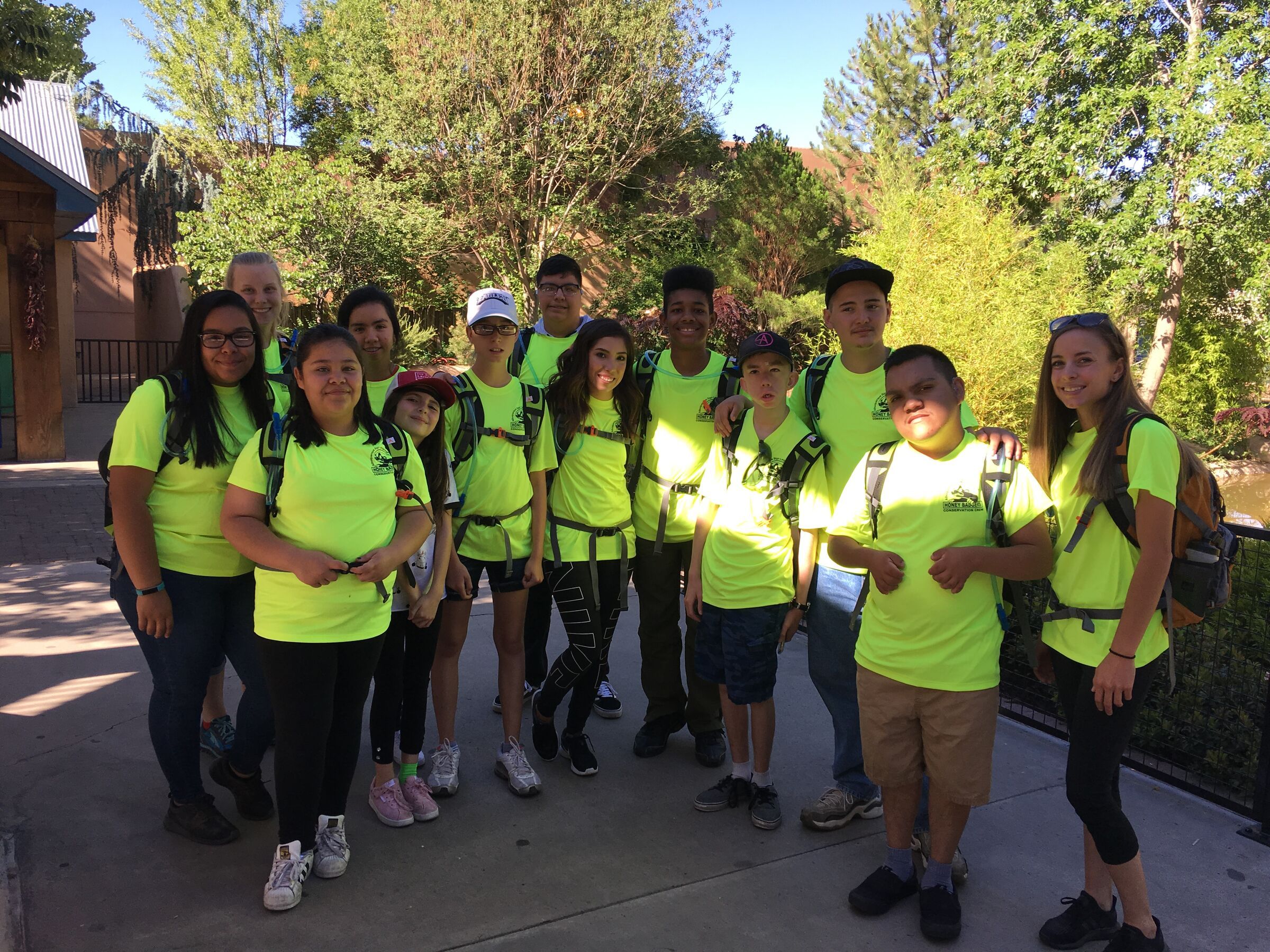 About twenty kids and leaders in matching bright yellow shirts and backpacks, smiling together under the trees.