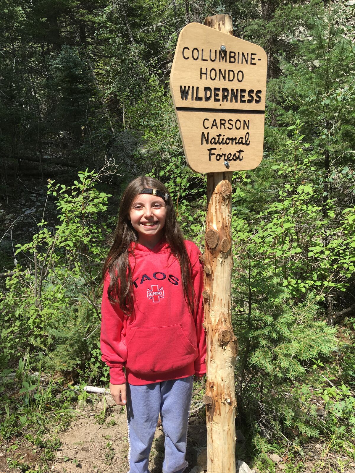 A smiling kid in a red Taos hoodie stands beside the carved Columbine-Hondo Wilderness sign.