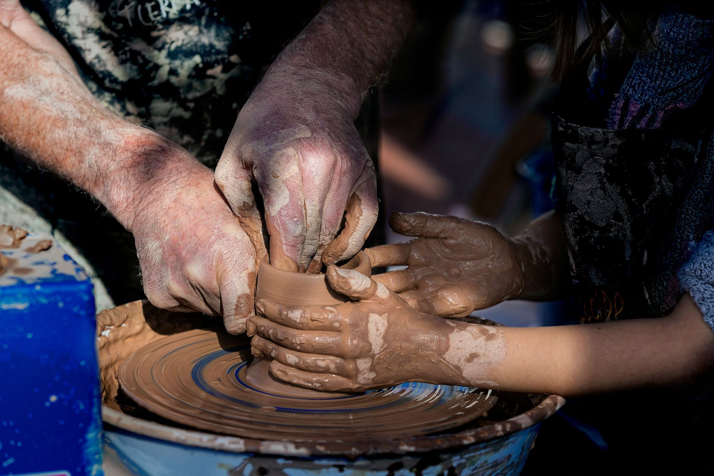 A potter's hands guide a child's as they shape a clay pot on the wheel.