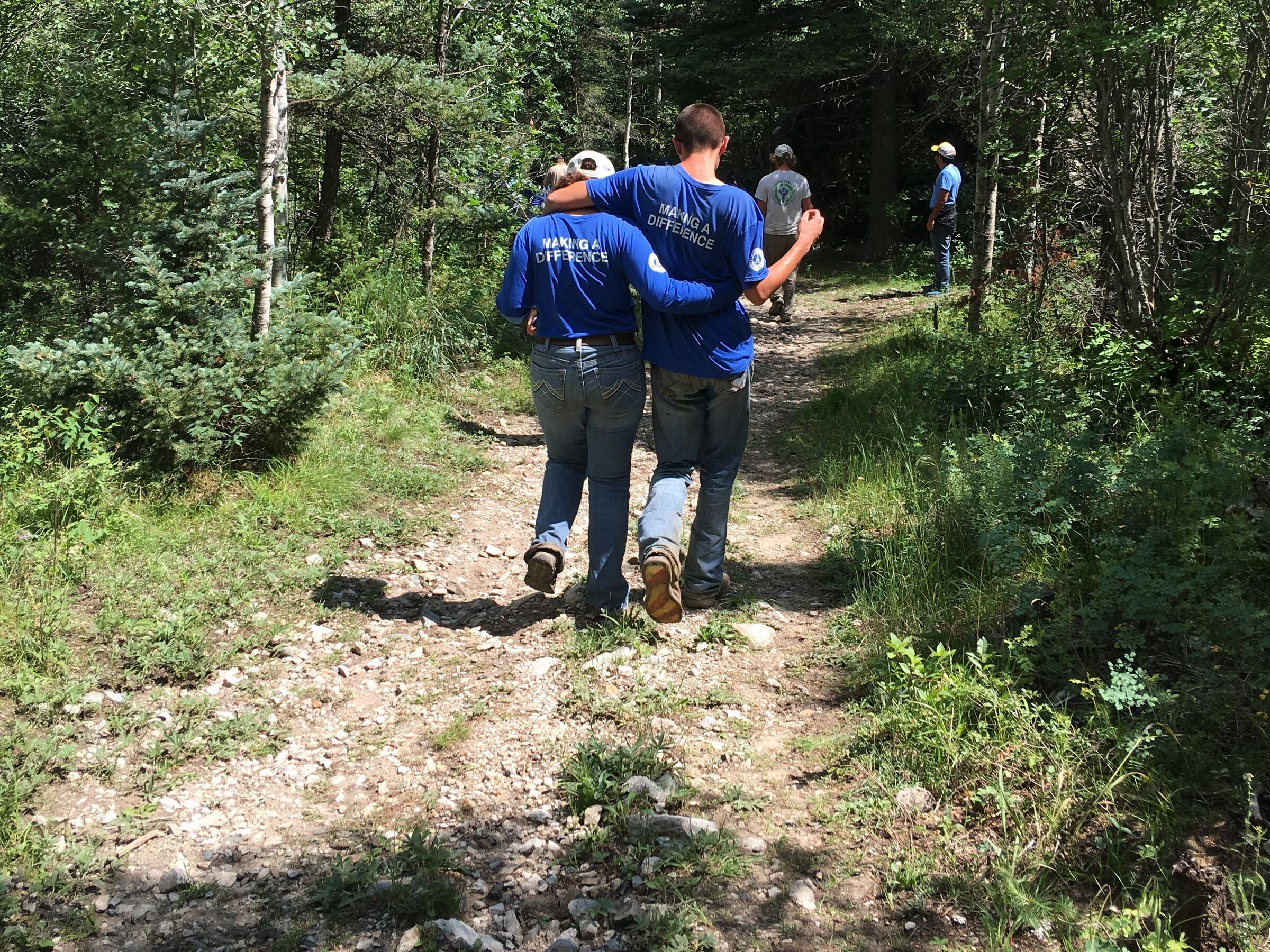 Dos personas de azul caminan por un sendero angosto hacia un bosque verde, de espaldas.